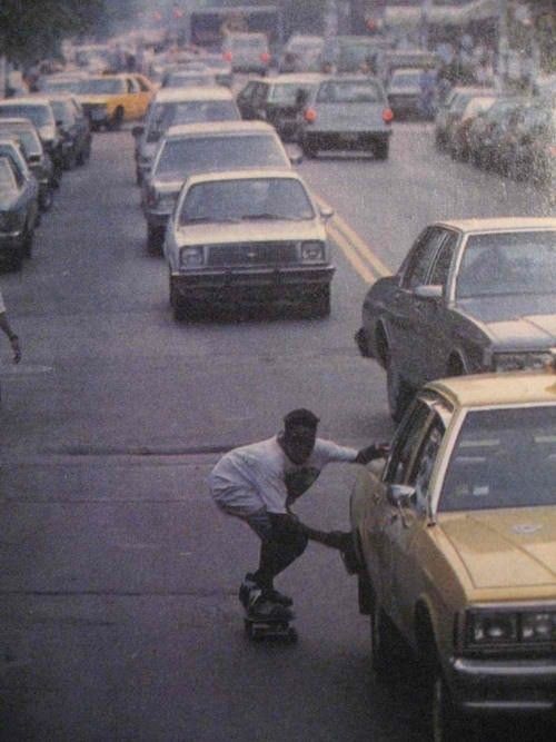 A skateboarder in a white t-shirt hitching a ride on the side of a yellow cab on a busy, traffic-filled city street lined with vintage vehicles.