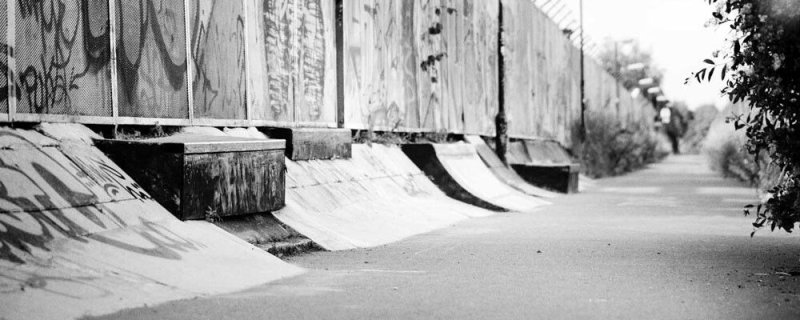 A black-and-white photograph of Train Bank  in Malmö an outdoor DIY skate spot featuring a paved pathway next to a long graffiti-covered fence with built-in concrete ramps, ledges, and quarterpipes.